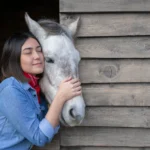 Horse owner gently embracing a white horse beside a wooden stable, illustrating the bond horse trust planning aims to protect.